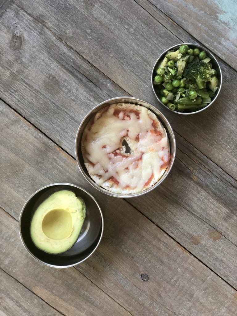 Pizza bagel with cheese, half of an avocado, mixed greens mix with broccoli, asparagus and peas